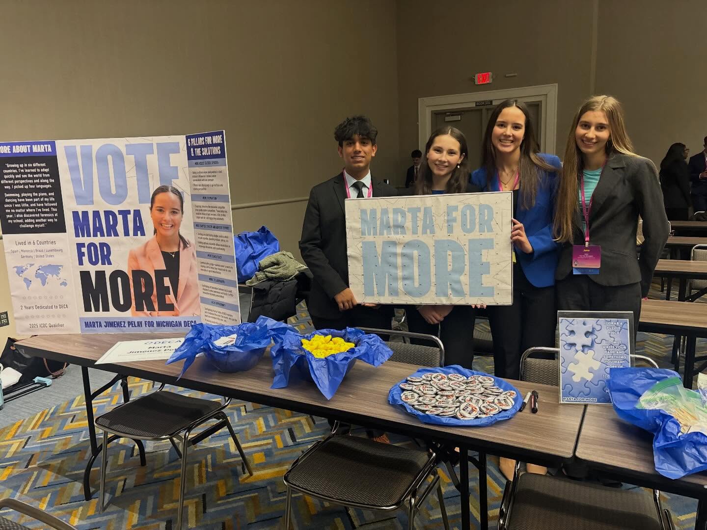 4 teenagers gathers around a both holding signs that say "Vote Marta for More". 