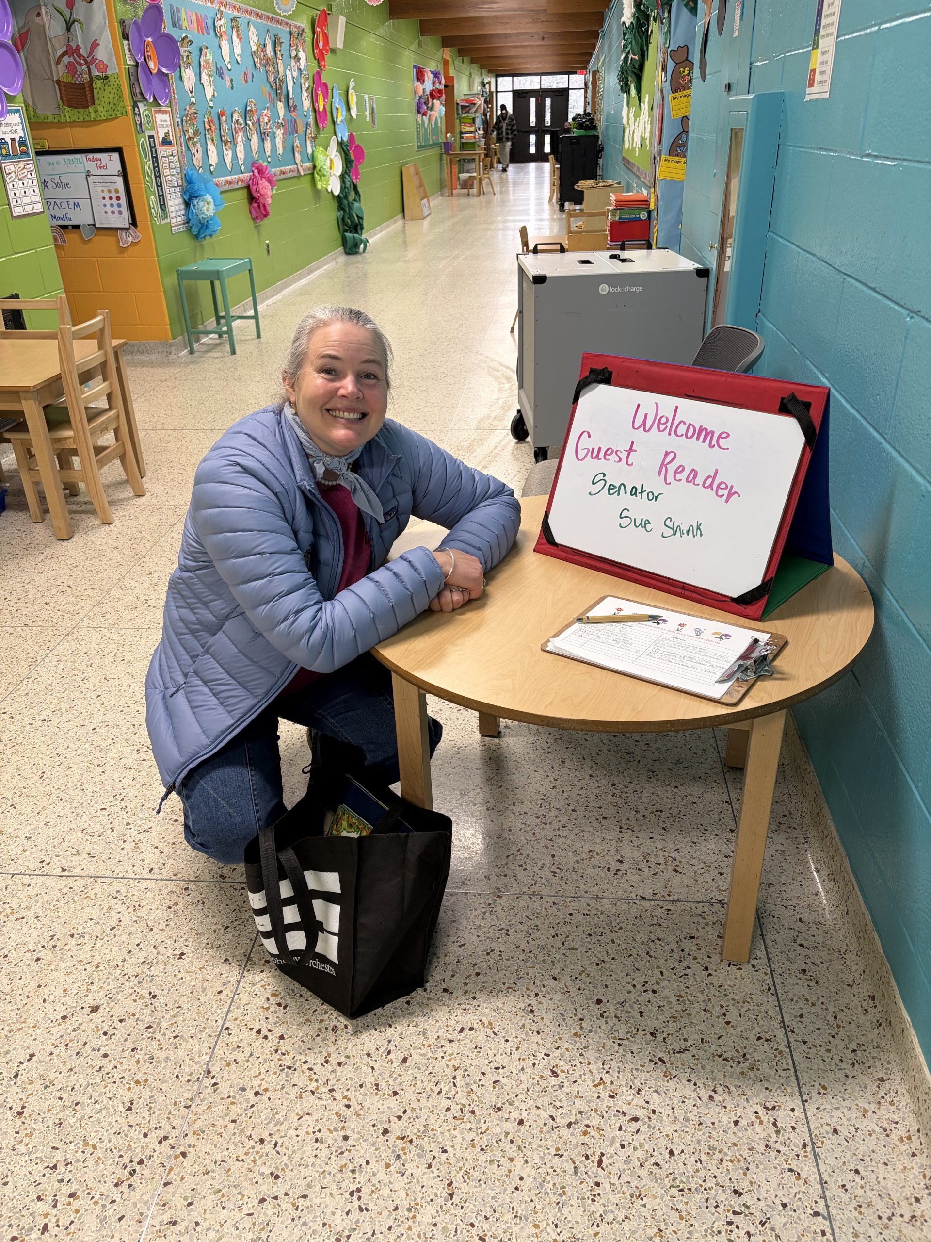 Sen Shink in an elementary school kneeling next to a sign that says "Welcome Guest Reader Senator Sue Shink"